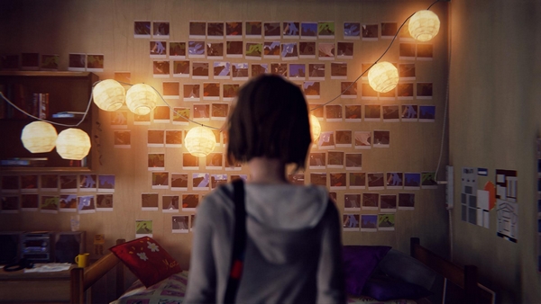A femme looking person standing in a bedroom. Wall with lots of photos on it. Lights hang across from the ceiling.